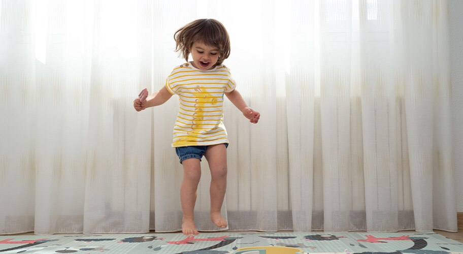toddler jumping on a colorful mat in front of curtains.
