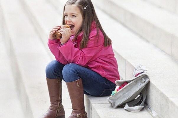 A little girl is sitting next to her backpack on the stairs, eating a sandwich.