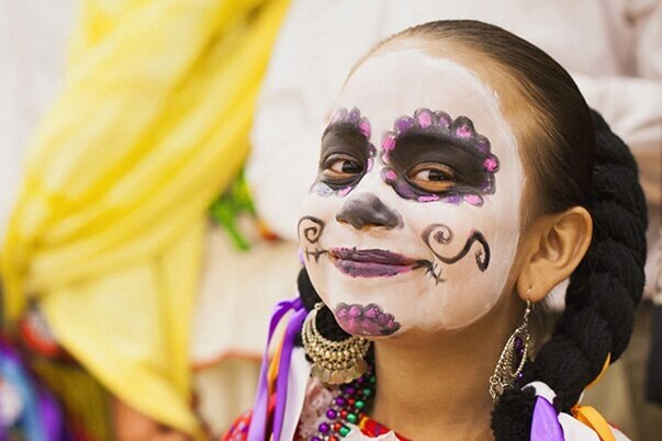 A little girl is made up and dressed to celebrate the Day of the Dead in front of a large group of celebrating people.