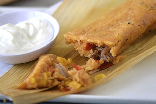 Cuban style tamales in corn husks on a white plate next to a bowl of sour cream.