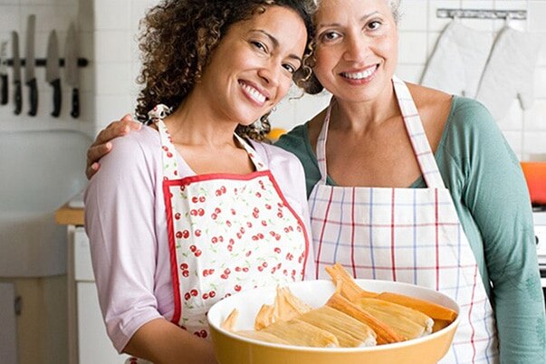 A woman and her mother smile as they pose with tamales they made in their kitchen.