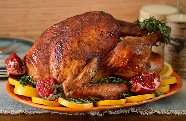 A whole roasted turkey on a bed of oranges, pomegranates, rosemary, and a large orange-colored plate.