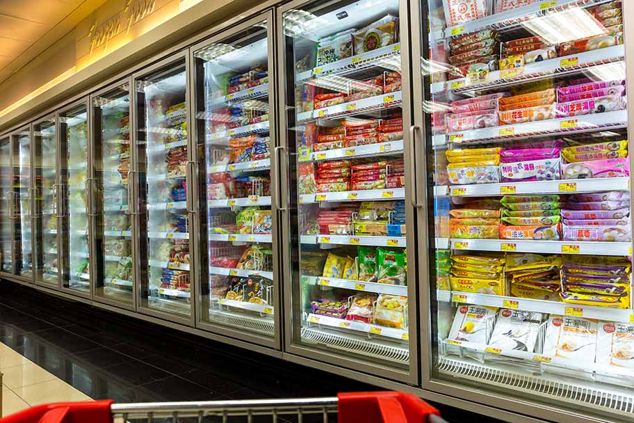 A shopping cart approaches large grocery store freezers filled with shelves of frozen food.