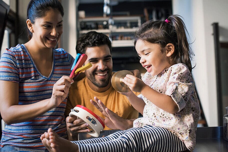 A toddler plays with cymbals as her parents cheer her on while playing a tambourine and noise maker.