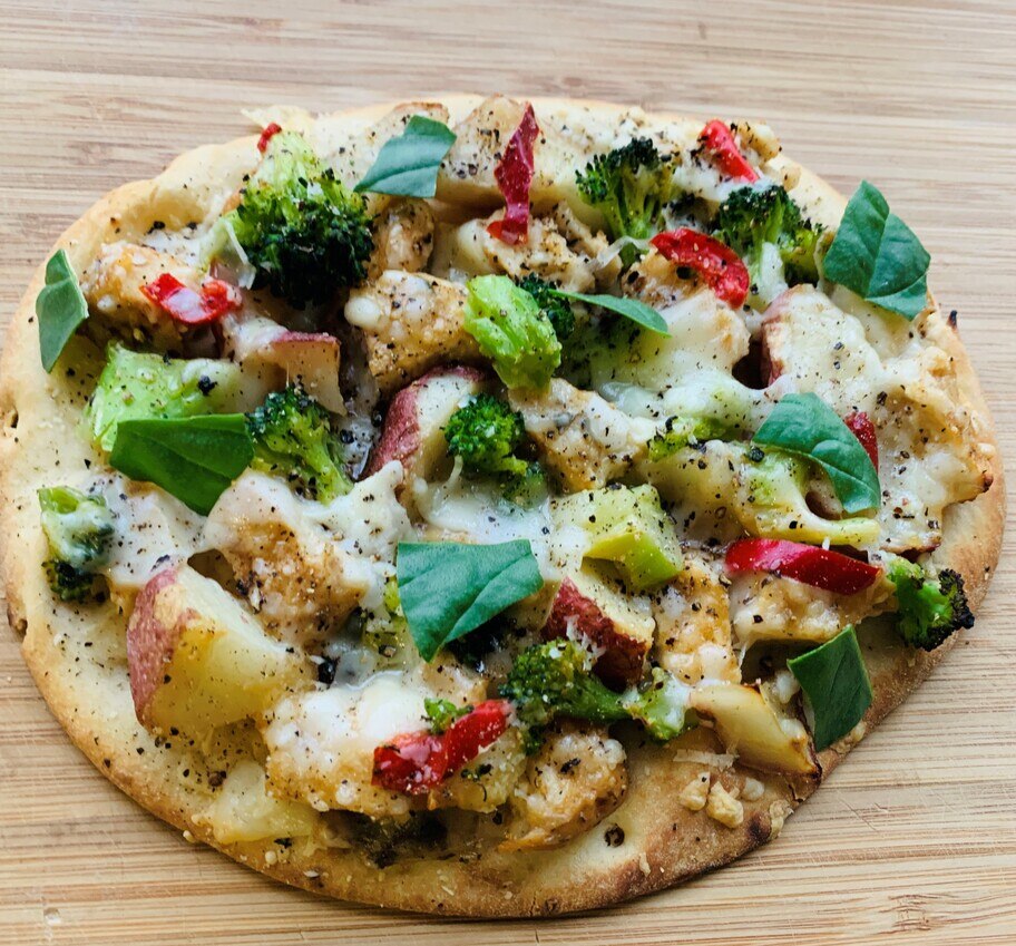 An herb roasted chicken flatbread with broccoli, basil and chicken all on a wooden counter. 