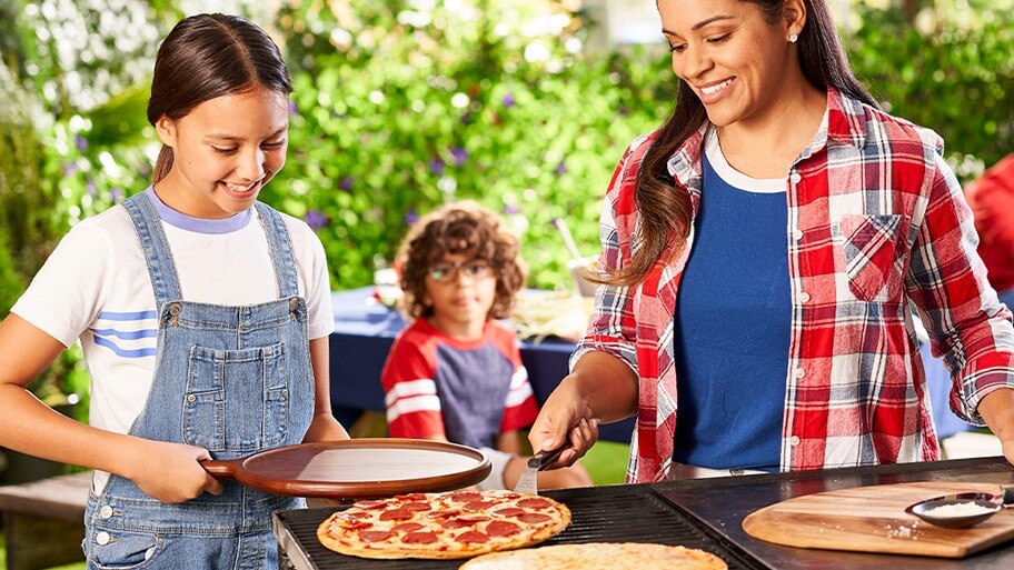 A mom lifts a pepperoni pizza off of a gas grill with a spatula as her son and daughter watch while holding a serving tray.d