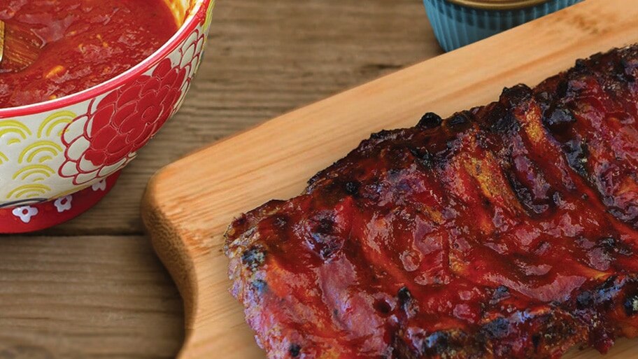A slab of ribs slathered in bbq sauce on a cutting board near a bowl of bbq sauce.