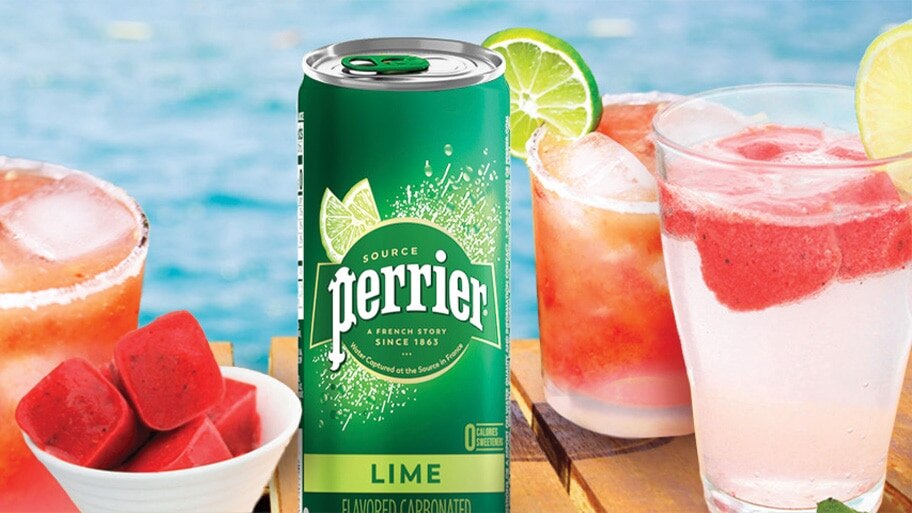 A can of lime Perrier by a bowl of watermelon ice cubes and glasses of Perrier fruit spritzers on a table by the pool.