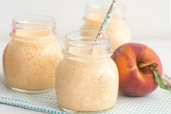 Three glass jars filled with peach smoothie beside a peach on a cloth napkin.