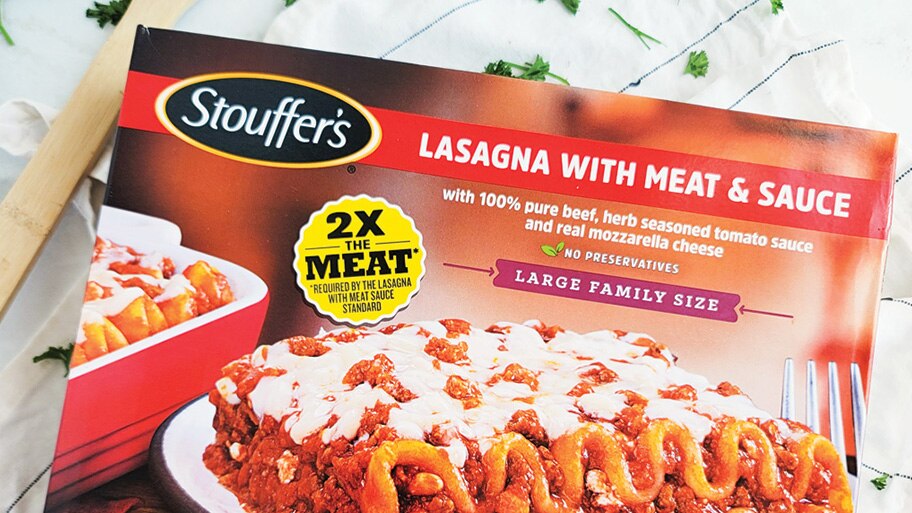 A box of Stouffer's lasagna with meat & sauce on a cloth napkin with scattered herbs.