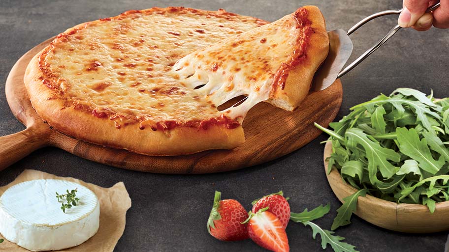 A DiGiorno pizza on a wood tray near a bowl of arugula, sliced strawberries, and a wheel of goat cheese.