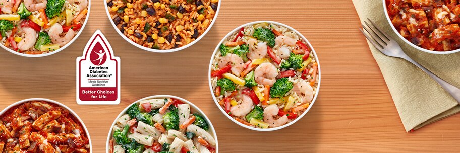 Overhead of Lean Cuisine balance bowls on wood table with forks, napkins, and American Diabetes Association badge.