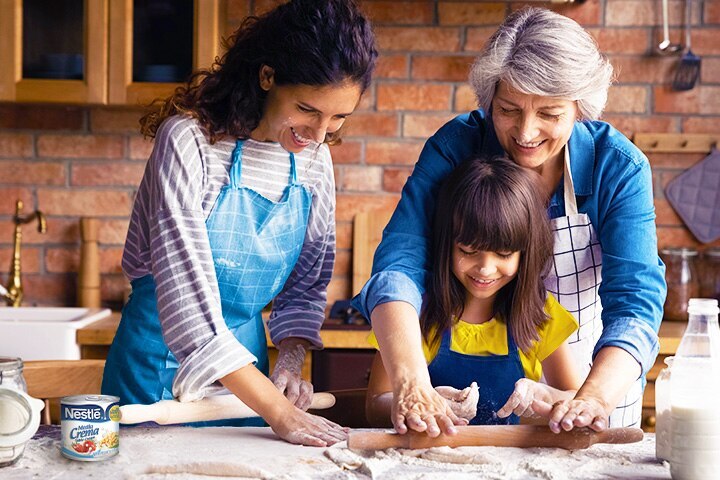 A mom and grandma help a young girl roll dough on a counter set with milk, flour and a can of Media Crema.