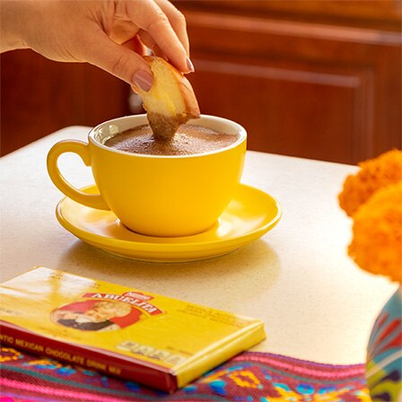 A hand dips a concha into a yellow mug of Mexican hot chocolate beside an Abuelita® bar.