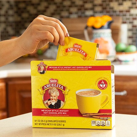 A hand pulls a packet of Abuelita® instant Mexican hot chocolate mix from the box.