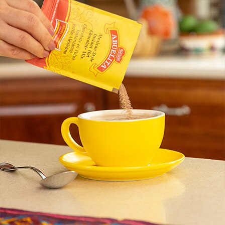A hand pours a packet of Abuelita® instant Mexican hot chocolate mix into a yellow mug.