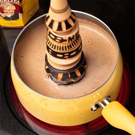 A decorative molinillo stirs Abuelita® Mexican hot chocolate in a yellow sauce pan.