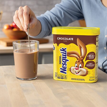 A hand stirs a glass of chocolate milk next to a canister of Nesquik chocolate flavored powder.