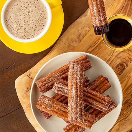 Un plato de churros sobre una tabla de cortar junto a una taza de Abuelita® y una taza de chocolate para mojar.