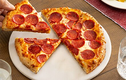 A hand, picking up a slice of pepperoni pizza from a white plate containing a whole pizza pie.