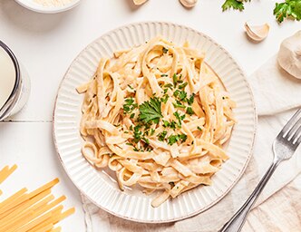 A plate of creamy alfredo surrounded by uncooked pasta, garlic and a fork.