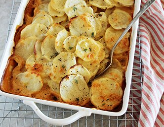 Un platillo con papas gratinadas y una cuchara sobre una rejilla y una toalla a rayas.