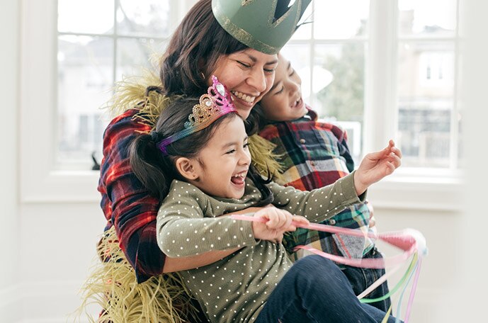 A mom laughs and hugs her children as they wear tiaras and crowns and play with magic wands.