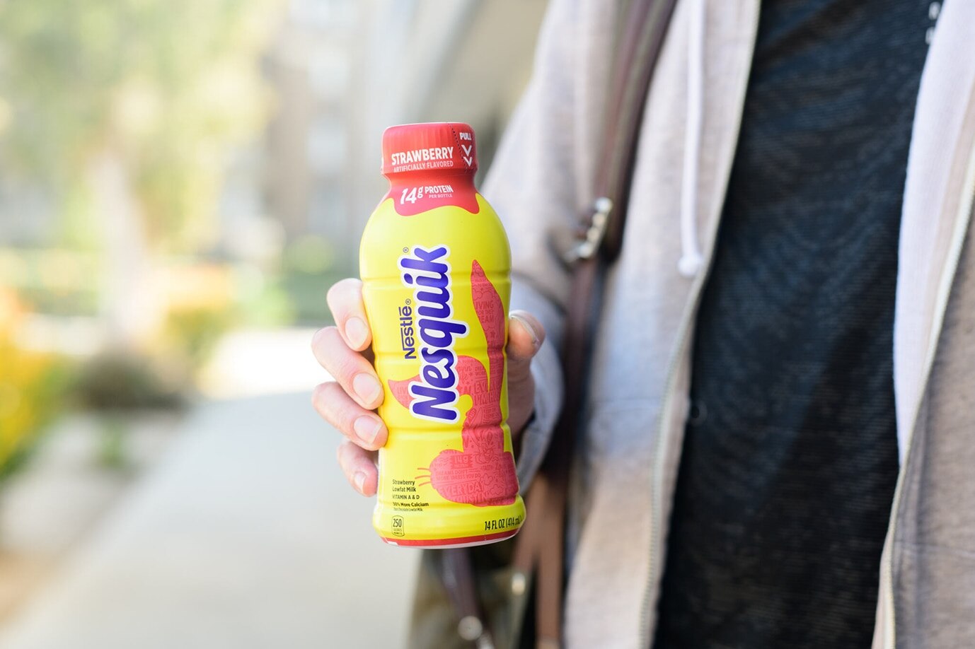 A person in a light gray hoodie holds a bottle of Nesquik Strawberry flavored milk outdoors.