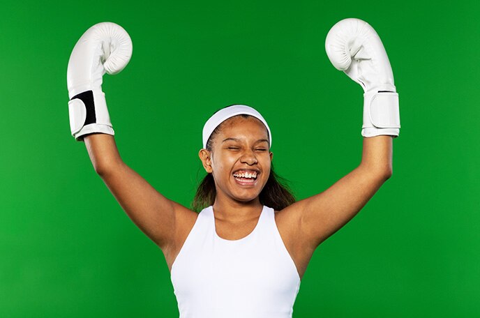 A teen girl in a white tank top, white headband and white boxing gloves lifts her arms and cheers.