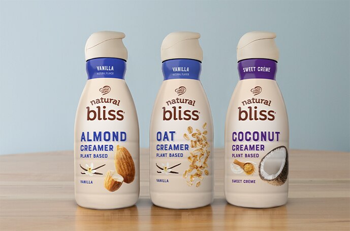 Three bottles of natural bliss plant based creamers lined up on a counter in front of a blue wall.