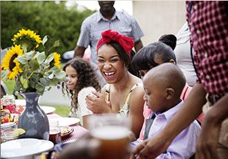 Una mujer joven sentada en una mesa de picnic sonriendo y riendo rodeada de su familia en una mesa servida