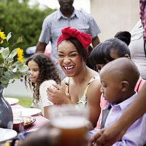A young woman sits at a picnic table smiling and laughing surrounded by her family and a table set with dinnerware and a red 