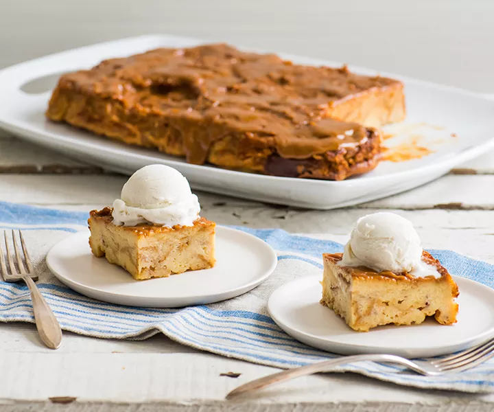 A white platter of bread pudding behind two plates with slices of tres leches bread pudding topped with vanilla ice cream.