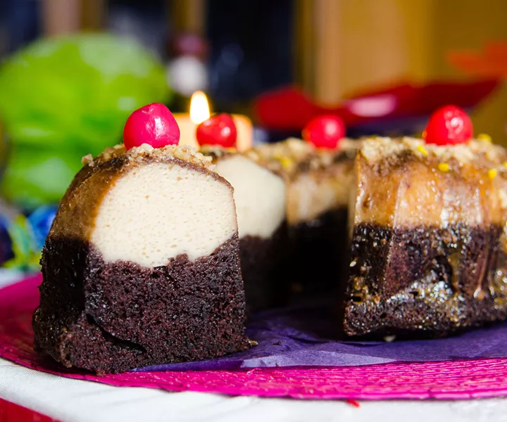 A layered flan cake with maraschino cherry garnish on a pink and purple cake stand. 