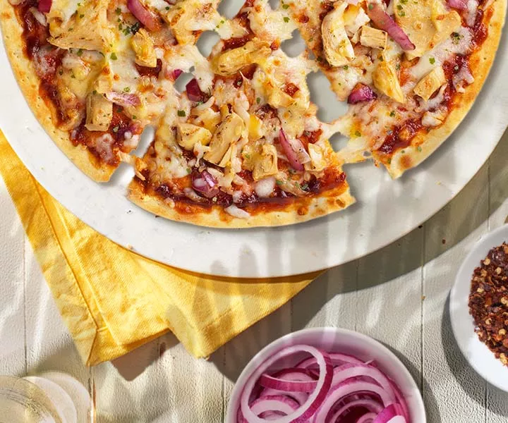A bbq recipe chicken pizza surrounded by bowls of red pepper and red onion slices and a glass of water.