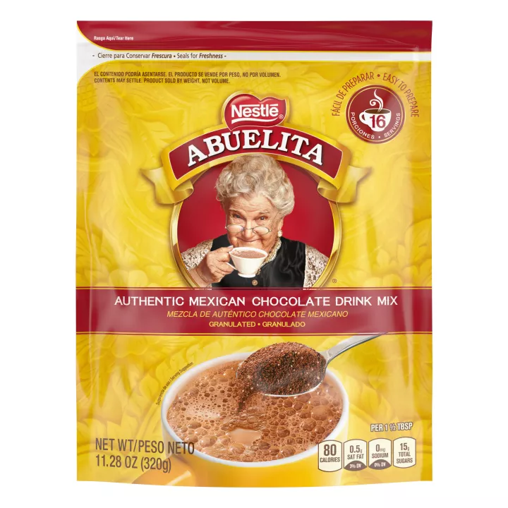 A yellow pouch with red labels and a spoon stirring hot chocolate mix into a yellow mug below the Abuelita logo.