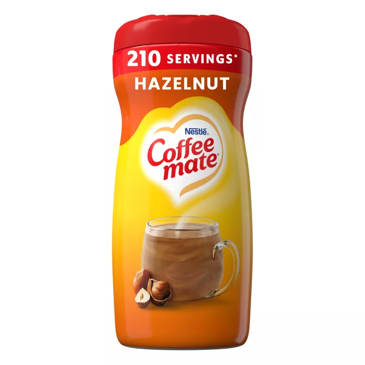 A yellow canister of Coffee mate Hazelnut Powder with a red label, mug of coffee, whole hazelnuts and the product logo.