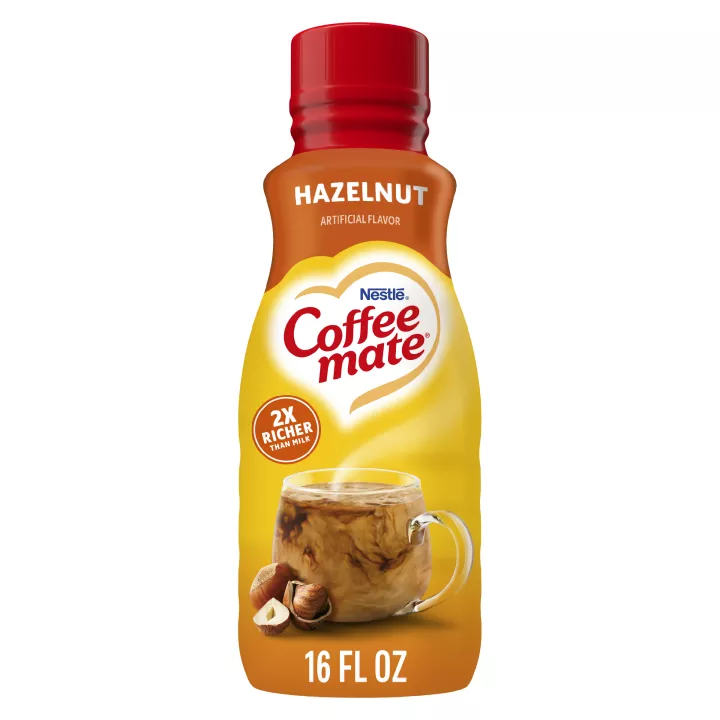 A bottle of Hazelnut Creamer with a red label  above a coffee mug, whole hazelnuts and the product logo.