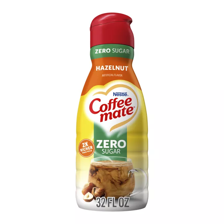 A yellow bottle of Zero Sugar Hazelnut Creamer with red & green labels above a coffee mug and whole hazelnuts.
