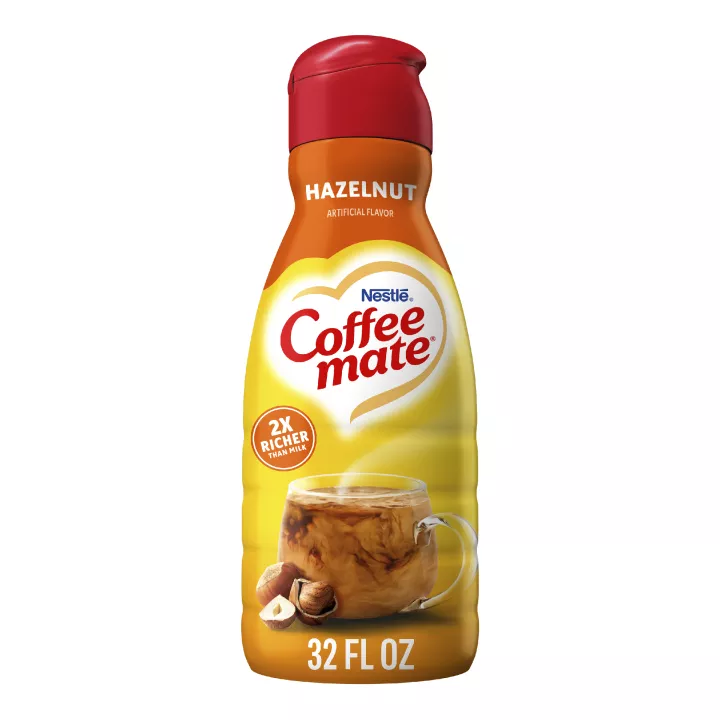 Una botella de creamer Hazelnut con una etiqueta roja encima de una taza de café, avellanas enteras y el logotipo del producto.