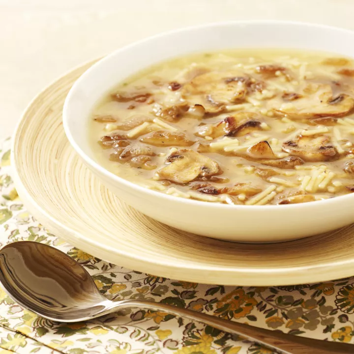 Foto con vista del ángulo izquierdo de un paquete de mezcla de Sopa de res con fideos MAGGI con una cuchara en un tazón de sopa de res con fideos encima de una etiqueta amarilla brillante con carne de res a término medio y cortada con cebollas y tomates debajo de la etiqueta amarilla.