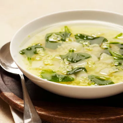 Foto con vista del ángulo derecho de un paquete de mezcla de Sopa sabor a pollo con arroz MAGGI con una cuchara en un tazón de sopa de arroz encima de una etiqueta amarilla brillante, y un pollo asado entero debajo de la etiqueta amarilla.