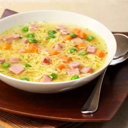 Foto con vista del ángulo izquierdo de un paquete de mezcla de Sopa sabor a pollo con fideos MAGGI con una cuchara en un tazón de sopa de fideos encima de una etiqueta amarilla brillante, y un pollo asado entero debajo de la etiqueta amarilla.