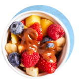 Close up of a fruit salad in a small white bowl with caramel drizzle on top, on a blue background.