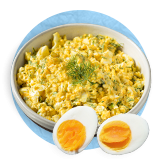 Close up of an egg salad in a white bowl and a hard boiled egg cut in half in front of bowl, on a blue background.