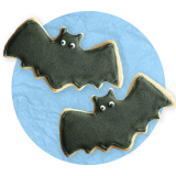 Close up of a pair of cookies shaped like flying bats with black icing and white eyes, on a blue background.
