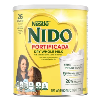 Una lata de Nido Fortificada con una etiqueta amarilla, una tapa blanca y un corazón dorado sobre un vaso de leche vertida.