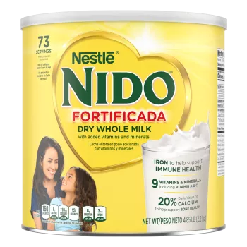 Una lata de Nido Fortificada con una etiqueta amarilla, una tapa blanca y un corazón dorado sobre un vaso de leche vertida.