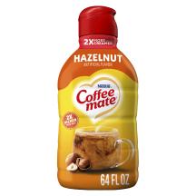 Una botella amarilla de creamer Coffee mate Hazelnut con una etiqueta roja encima de una taza de café, avellanas enteras y el logotipo del producto.