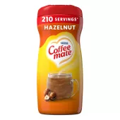 Una lata amarilla de creamer en polvo Coffee mate Hazelnut con una etiqueta roja, una taza de café, avellanas enteras y el logotipo del producto.